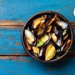 Mussels in clay bowl, glass of white wine and lemon on wooden blue background