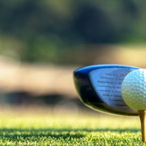 Close up golf ball and driver, player doing golf swing tee off on the green sunset evening time, presumably does exercise. Healthy and Lifestyle Concept.