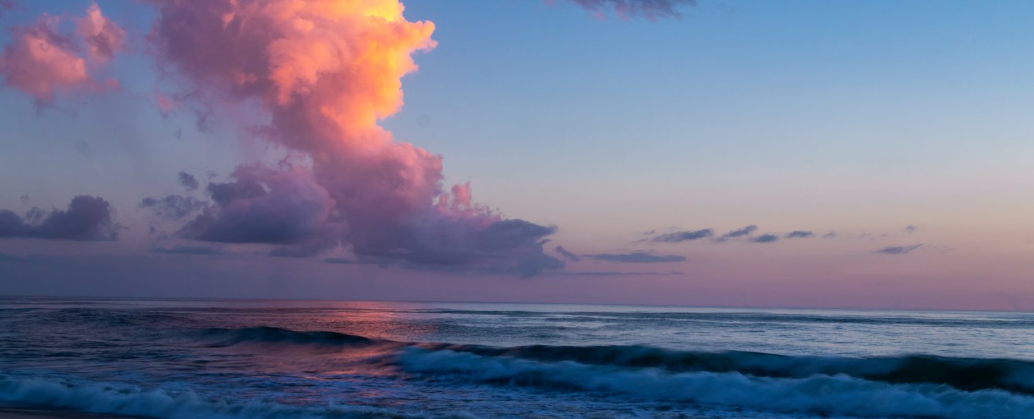 Amazing tall clouds over the ocean reflect the colors of the morning sun as it breaks over the horizon. Shades of purple, pink and blue in the sky and reflected on the water.