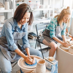 Two girls friends smiling and talking while working on potters wheel making clay handmade craft in pottery workshop, friendship and guidance concept