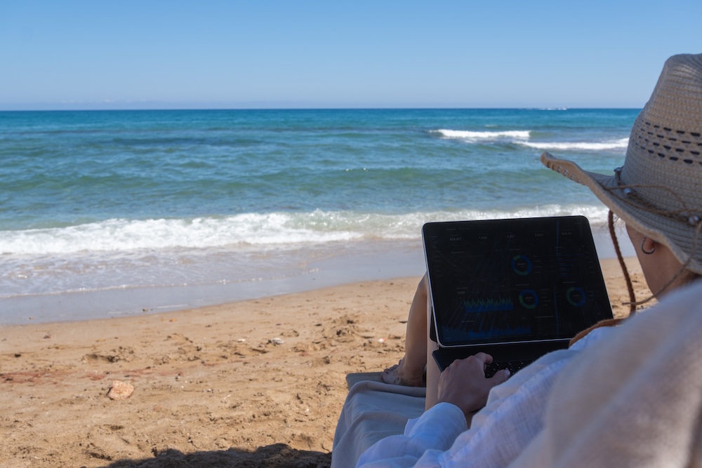 Woman in a straw hat sits on a beach lounge chair and works on a tablet with financial charts against a blue ocean background.