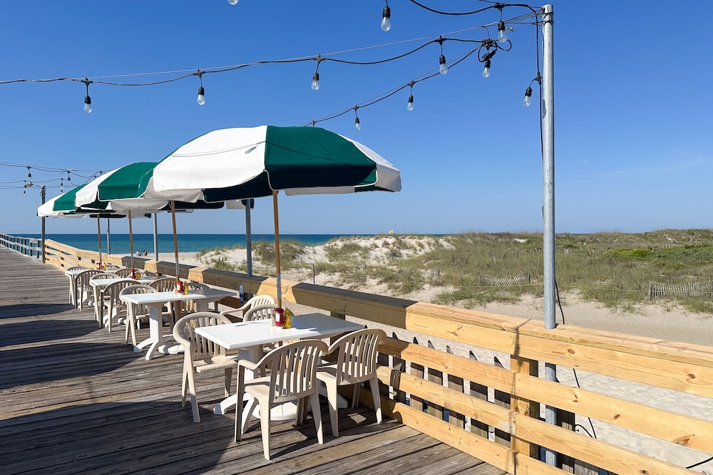 Outdoor dining tables with striped umbrellas overlook sandy dunes and the Atlantic Ocean from a beachfront boardwalk pier in Atlantic Beach, North Carolina on a sunny summer day.